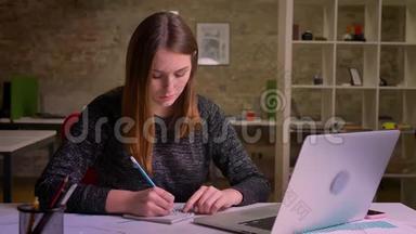 年轻的红头发妇女在笔记本电脑前工作，在办公室里用钢笔和笔记本做笔记